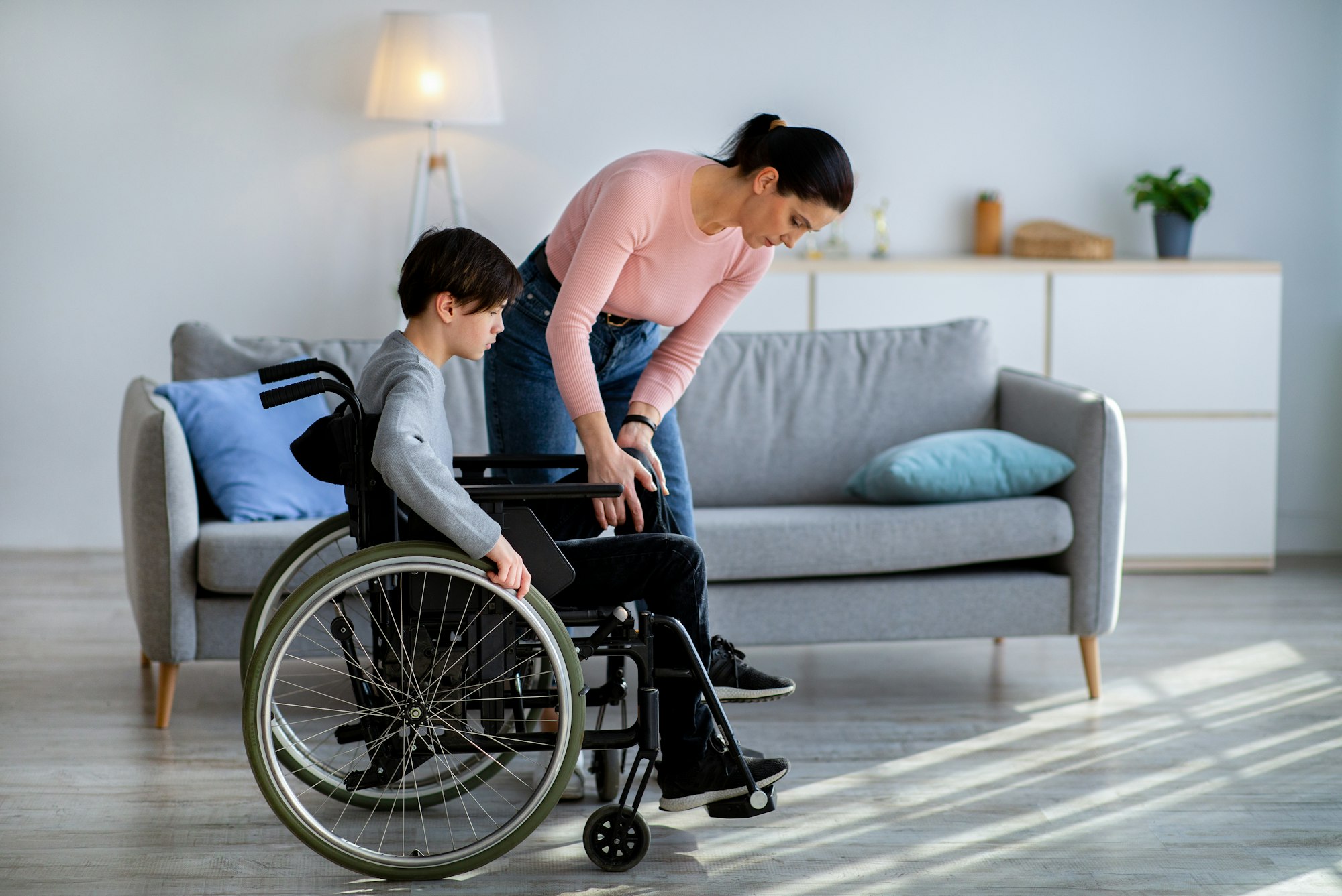 Young mother helping her disabled teen son in wheelchair move his leg at home, copy space